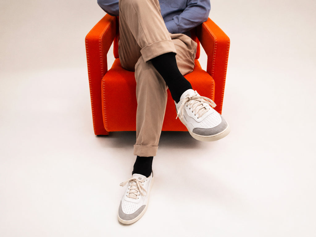 A person seated in an orange armchair wearing beige pants, black socks, and white low-top sneakers with gray toe panels, photographed against a clean light gray studio background.