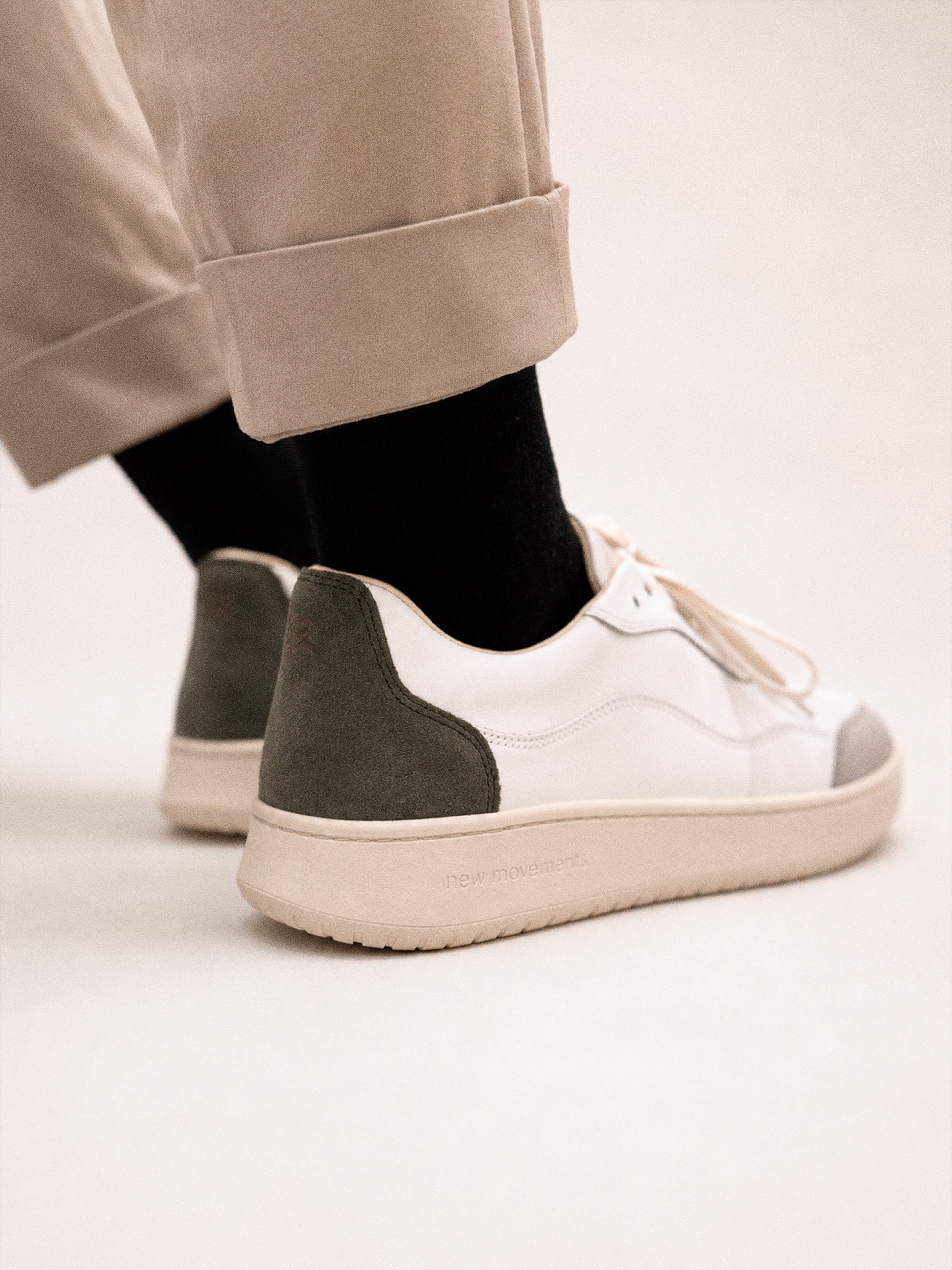 White low-top sneakers with dark olive green suede heel panels and beige soles, shown on a person wearing beige trousers and black socks against a light studio background.