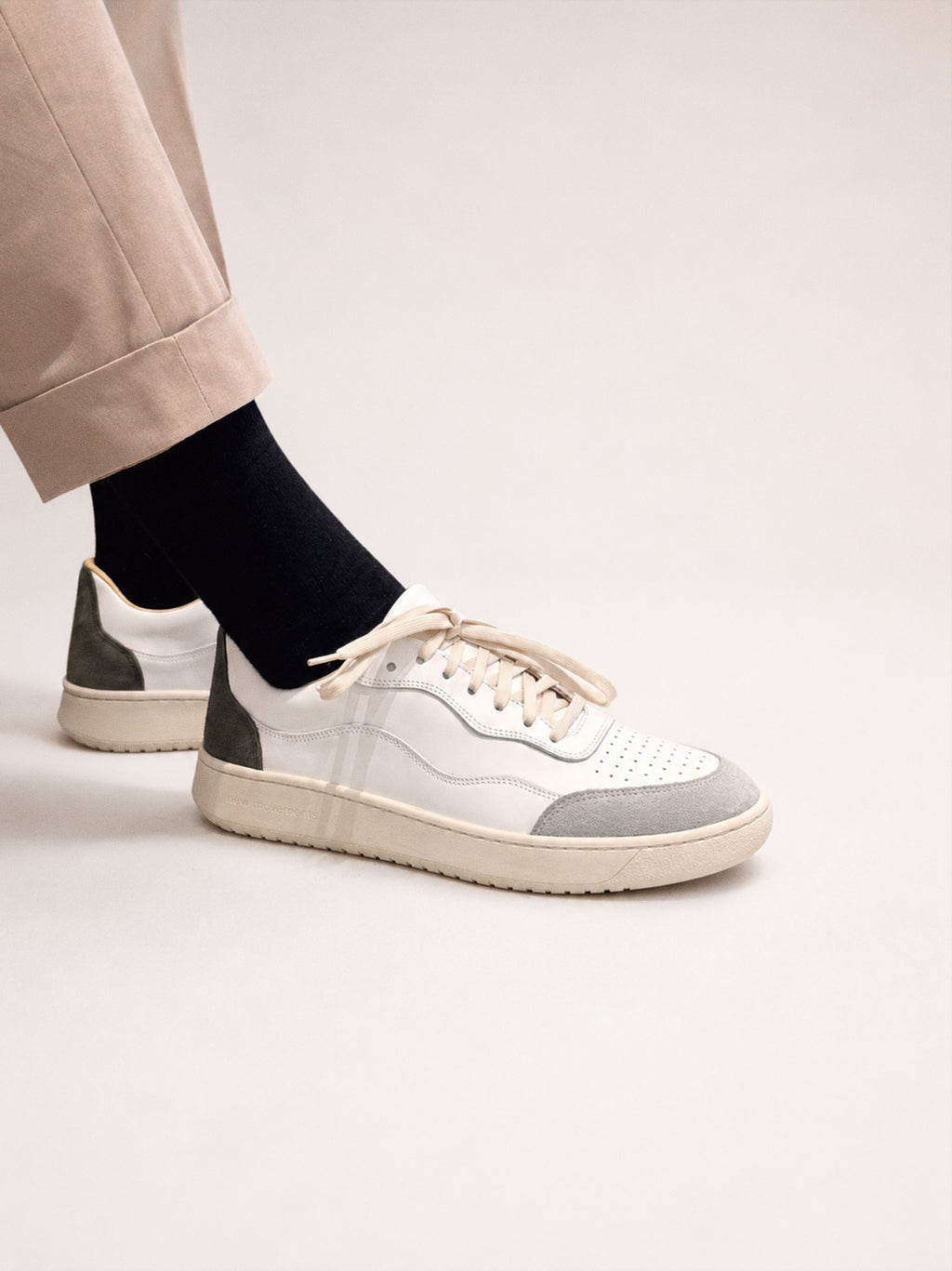 A pair of low-top sneakers in white, light gray, and dark green, shown on a model wearing beige trousers and black socks against a light beige studio background.