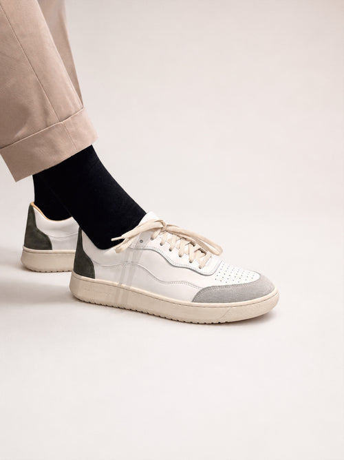 A pair of low-top sneakers in white, light gray, and dark green, shown on a model wearing beige trousers and black socks against a light beige studio background.